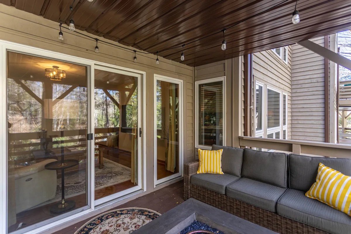 This image features a comfortable outdoor seating area with a sofa adorned with yellow striped pillows, glass doors leading into the house, a decorative rug, and hanging light fixtures. The space is set for relaxation or gathering, with hints of both indoor and outdoor ambiance.