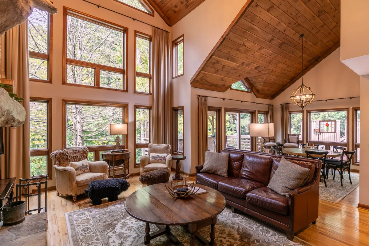 A bright, open-concept living and dining area with rustic wooden ceilings, elegant decor, a spacious seating area, and large windows that flood the room with natural light. The adjacent dining table is set for shared meals, and nature views create a serene backdrop.