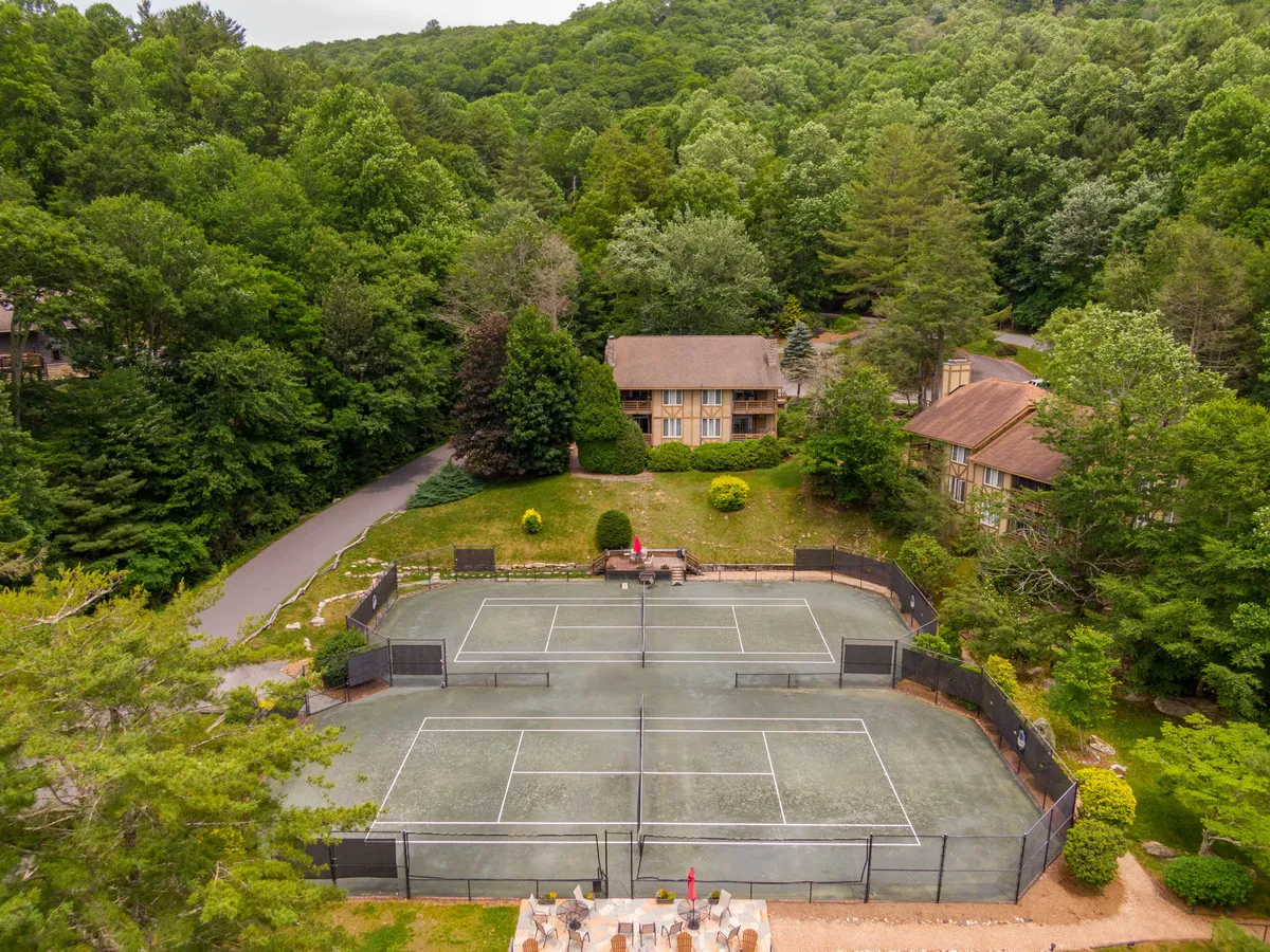A wide-angle view of the Yonahlossee property with tennis courts in the foreground, surrounded by lush greenery and several main and auxiliary buildings. The courts are active, inviting, and set in a peaceful mountain environment, reinforcing a sense of recreation and community.