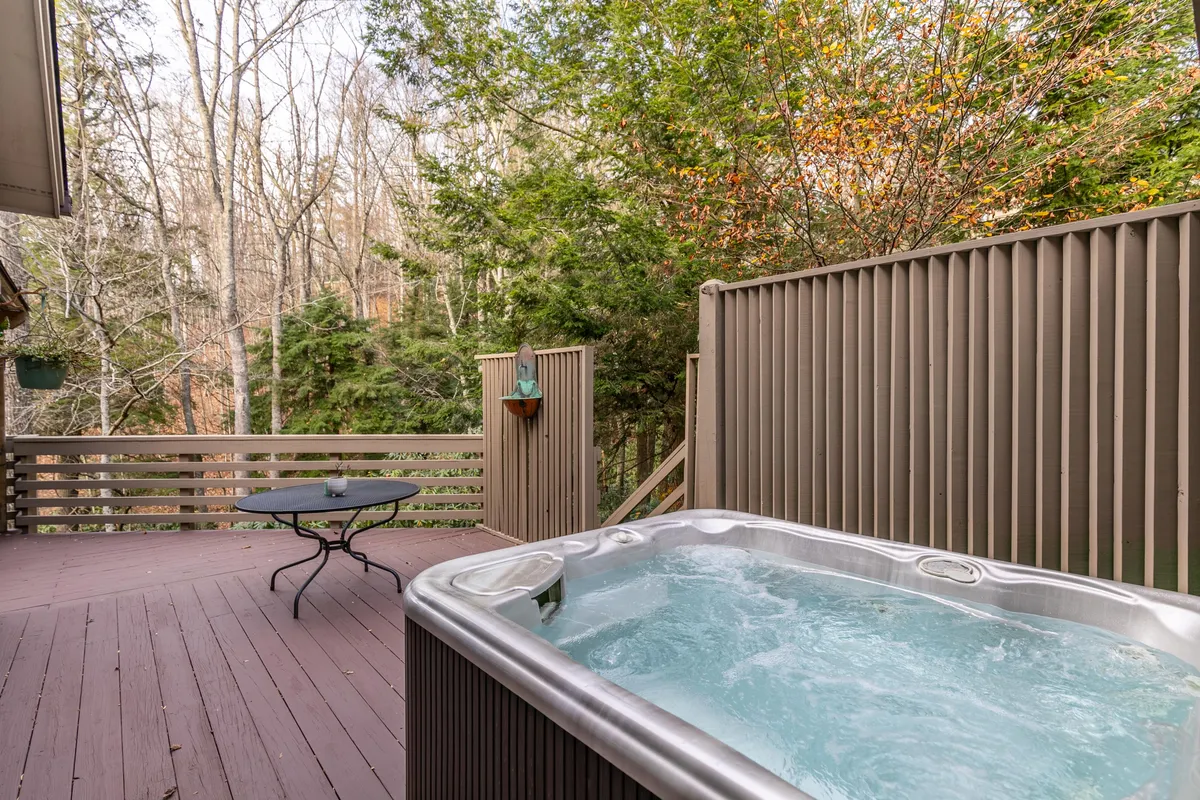 This exterior shot highlights the outdoor hot tub area, set among trees with a nearby outdoor table. The tranquil scene hints at relaxing soaks after cold-weather adventures, and the natural setting reinforces the connection to the mountain environment.