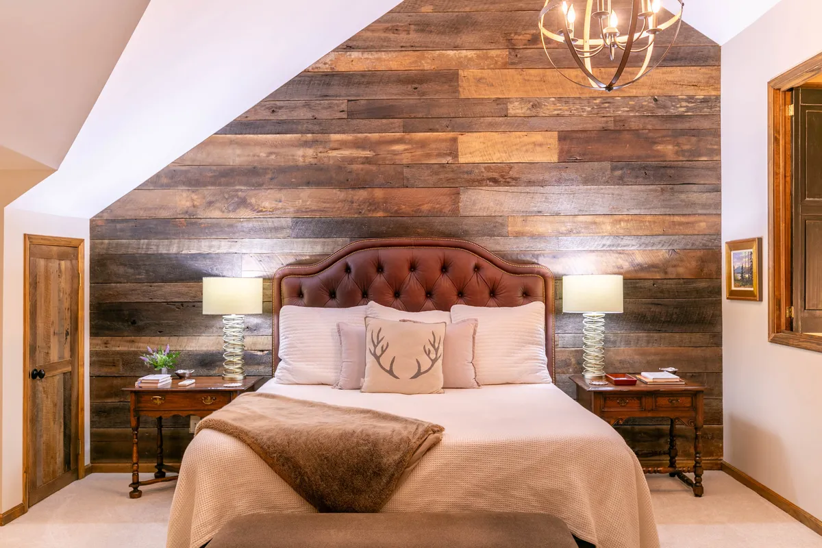 A bedroom with a wooden accent wall, an upholstered headboard, and stylish lamps on either side of the bed. Soft, decorative pillows add warmth, and the overall palette is cozy and inviting—a space designed for relaxation and rejuvenation.
