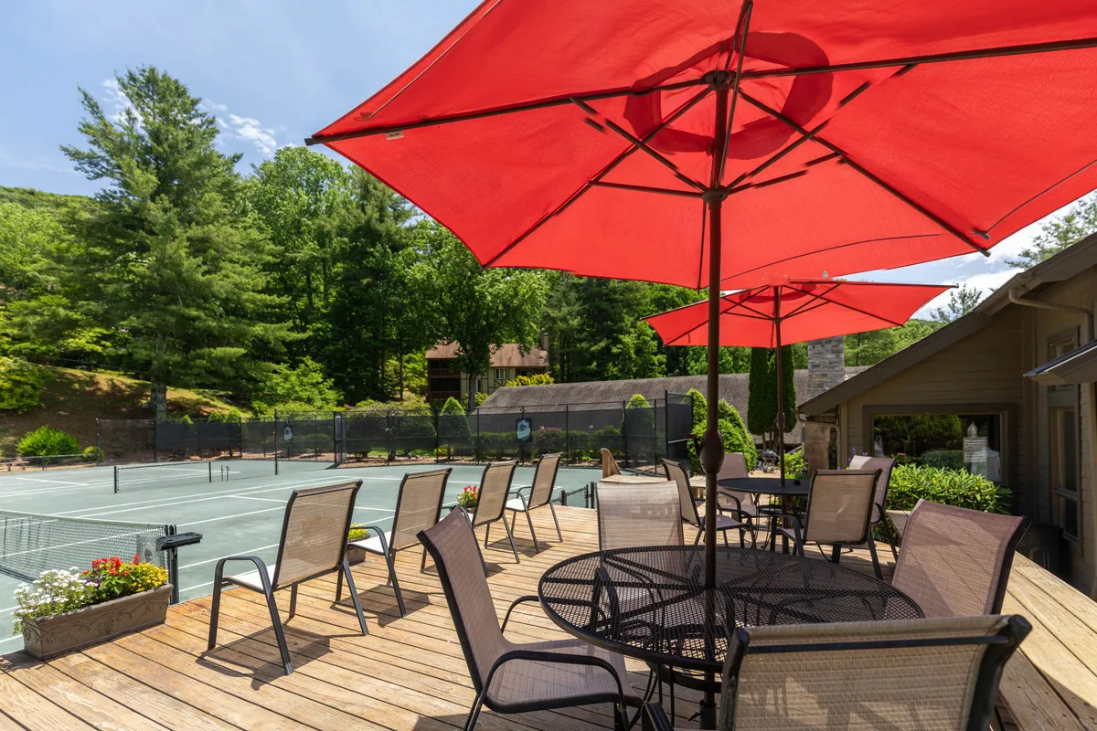 A vibrant exterior view of the Yonahlossee Racquet Club featuring outdoor clay tennis courts surrounded by red umbrellas, a wooden deck, and seating where guests are gathered. The scene is lively with people playing tennis, relaxing outdoors, and engaging in social activities under sunny skies.