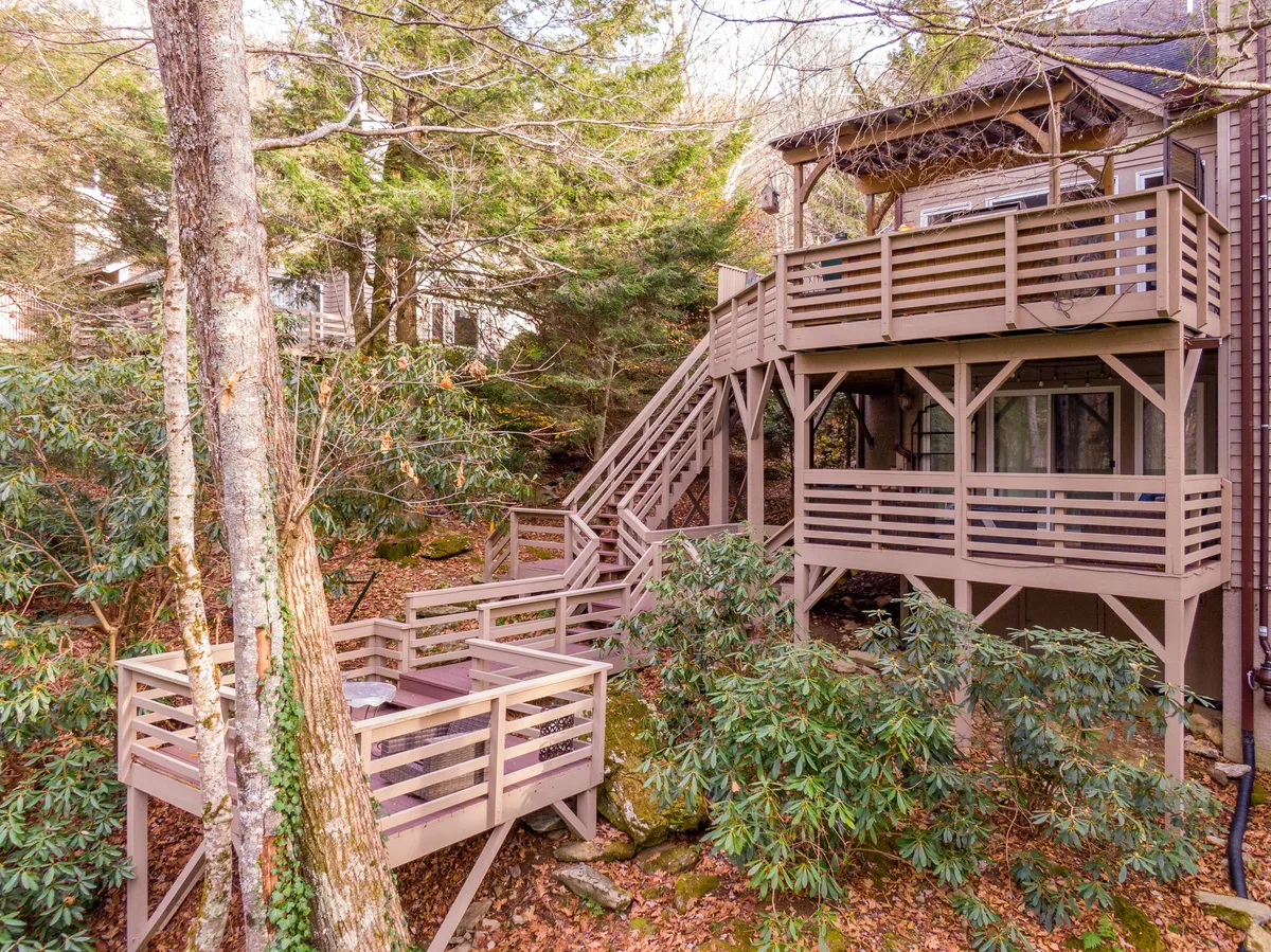 The image showcases the exterior of a luxury townhome nestled among lush trees, with a spacious multi-level deck featuring wooden railings that overlook a rocky, wooded landscape. The peaceful setting suggests opportunities for outdoor relaxation, family gatherings, and enjoying the natural beauty that surrounds the property.