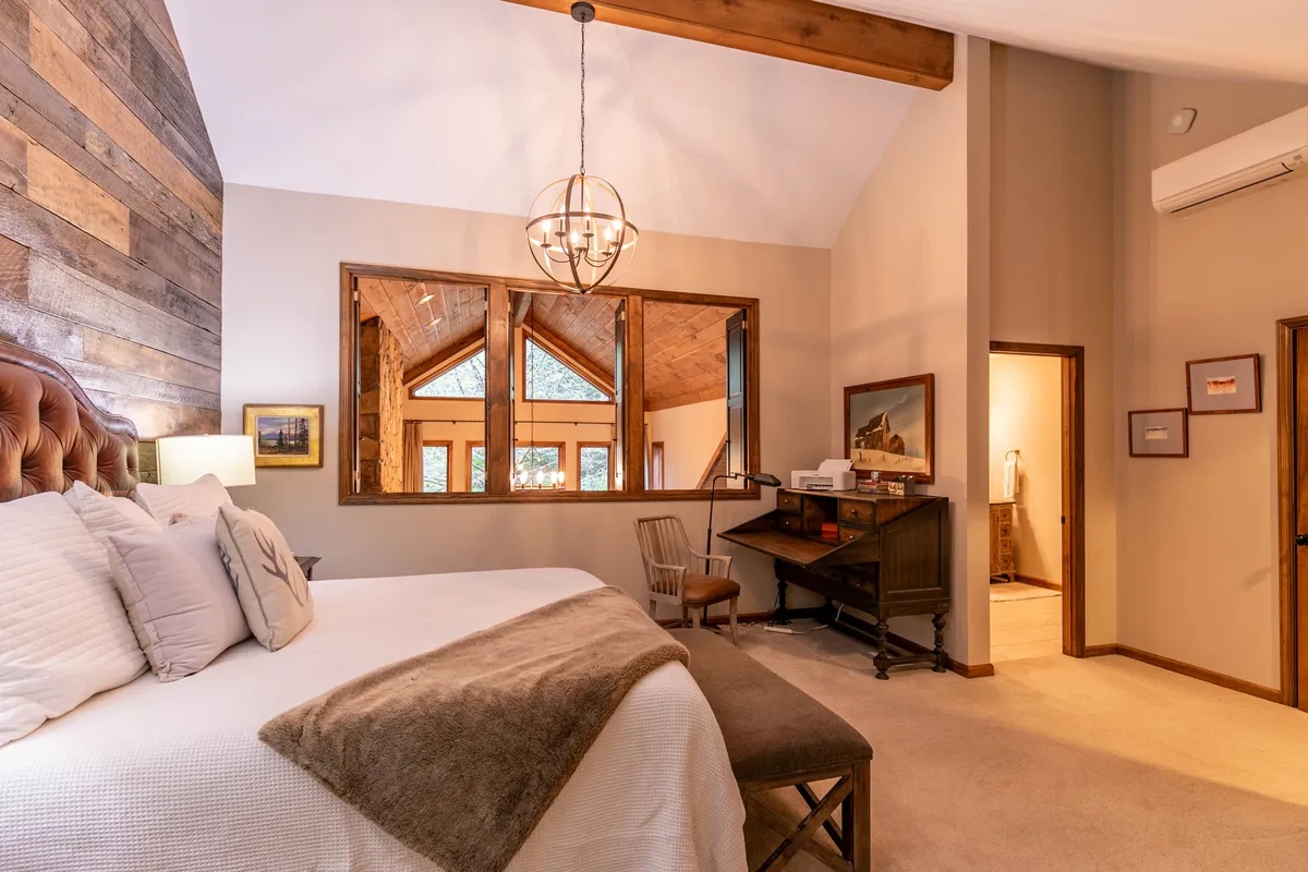 A tranquil bedroom featuring a plush bed with soft bedding and a throw, a large window streaming in natural light, a chandelier overhead, and a wooden desk and chair tucked by the window. The room feels airy and private, with a refined yet restful style ideal for a primary or master suite.