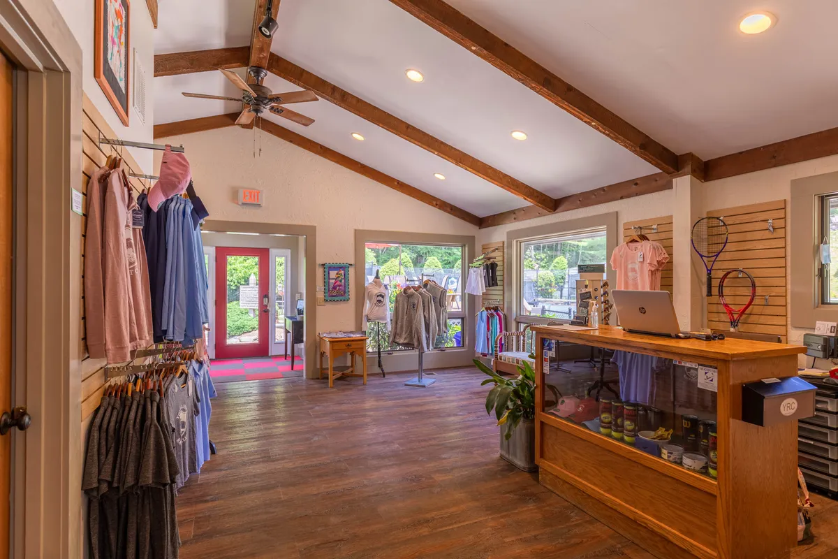 Inside a boutique shop, clothing racks, a display table, and a cash register are illuminated by large windows, creating a bright and inviting shopping atmosphere. The scene suggests browsing unique items and engaging with friendly staff.