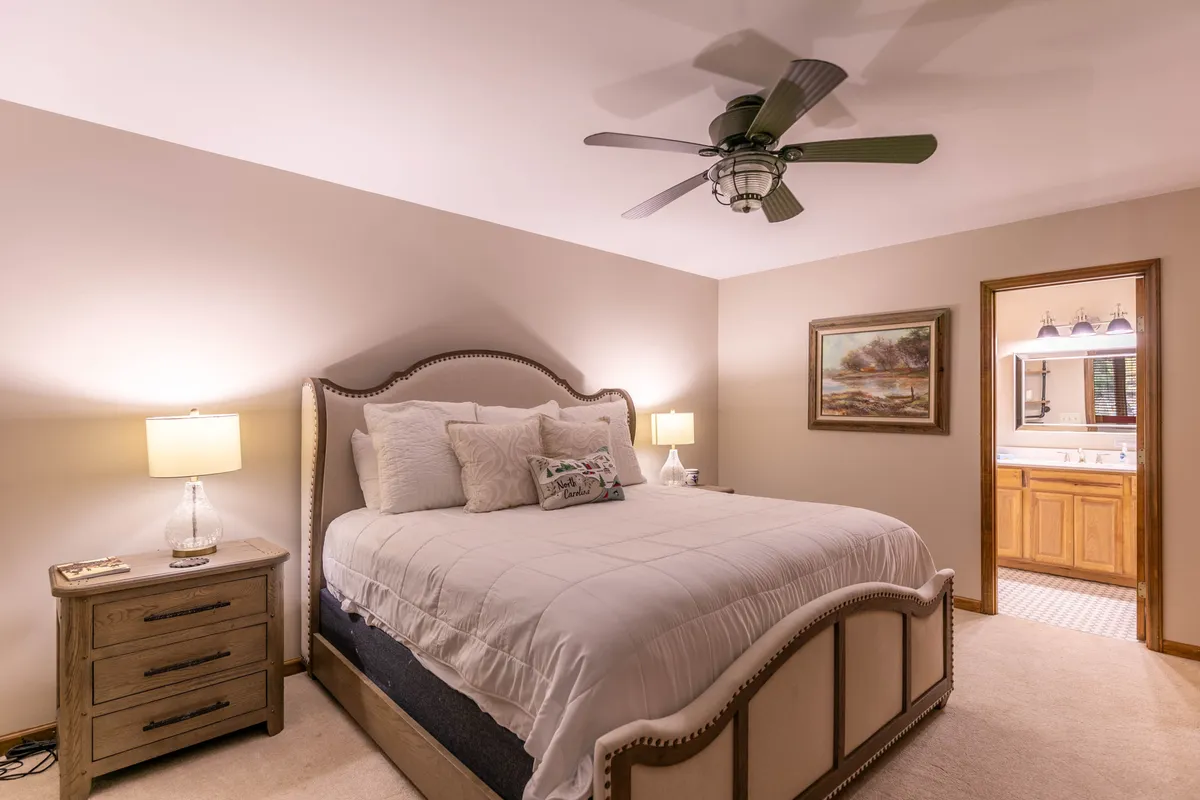A welcoming bedroom with a large bed, nightstands and lamps on either side, a painting above the headboard, and a doorway that leads to an ensuite bathroom. The space feels peaceful, organized, and restful.