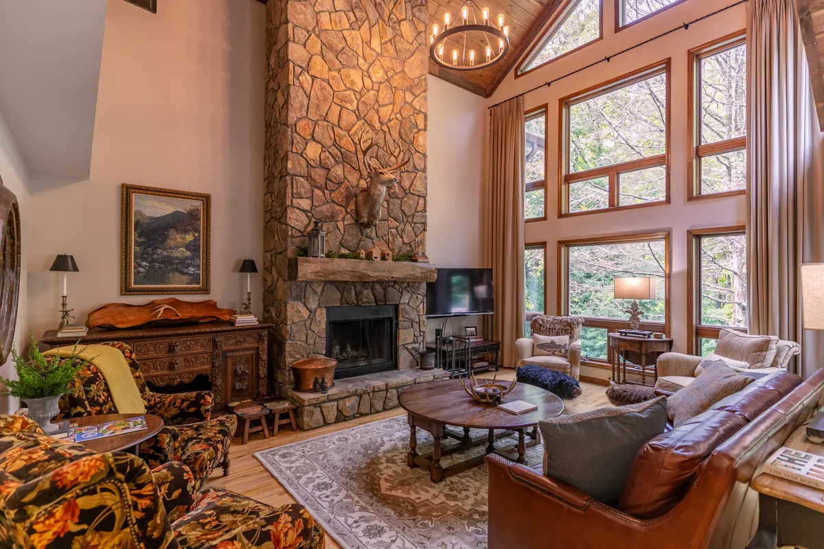 This image captures the great room, featuring a dramatic floor-to-ceiling stone fireplace, expansive two-story windows framing a view of nature, a plush seating arrangement, and a striking chandelier overhead. Sunlight pours in, creating an inviting, airy atmosphere. The cozy sofa and tasteful artwork enhance the space’s comfort and style.