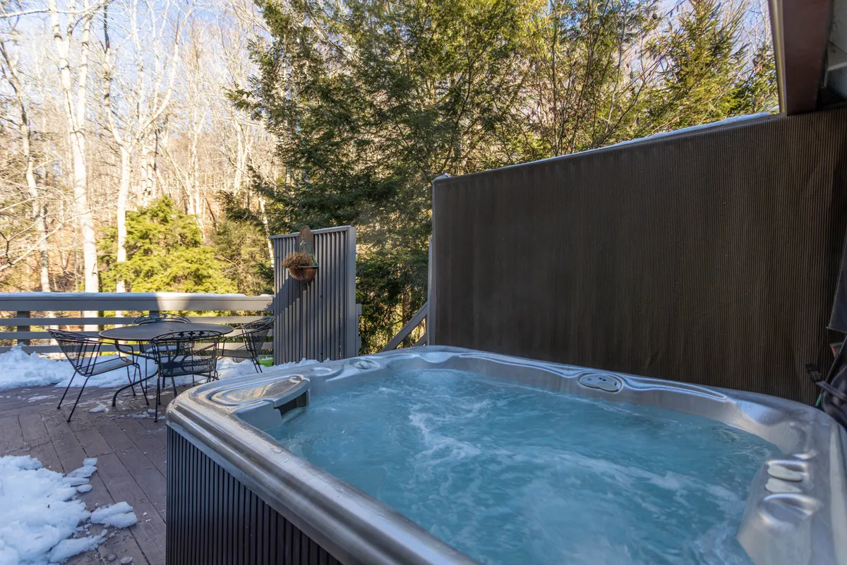 This image shows a spacious outdoor deck nestled among tall trees, featuring a bubbling hot tub and a comfortable seating area with a table—all surrounded by natural greenery. The ambiance is tranquil, inviting guests to relax outdoors, whether soaking in the hot tub or enjoying a meal al fresco.